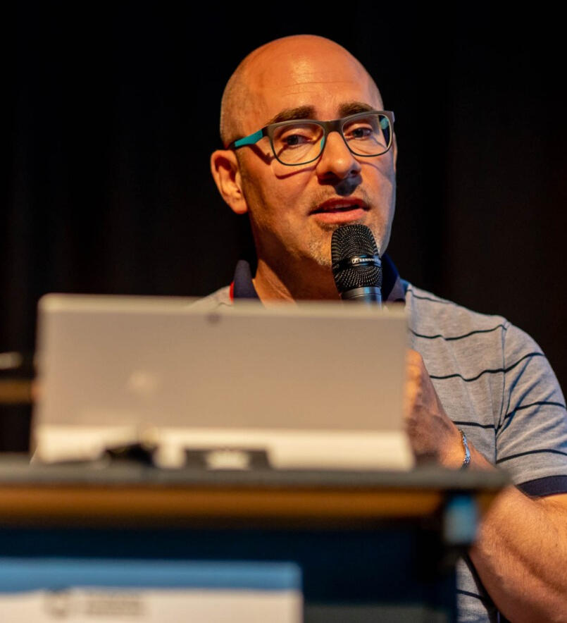 Matthias Blaß as Speaker on a conference. He holds a microphone behind a notebook talking to the crowd.